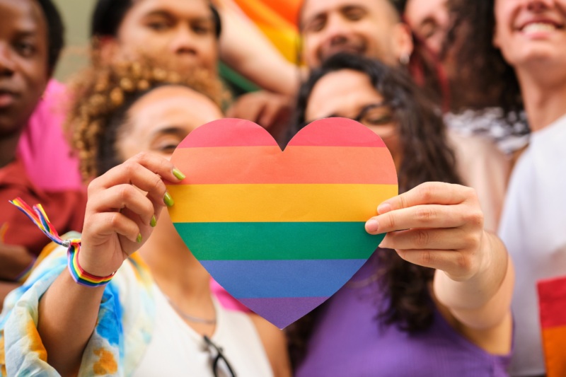 Close-up of two individuals holding a heart-shaped paper cutout with rainbow stripes, with a blurred crowd of people in the background.