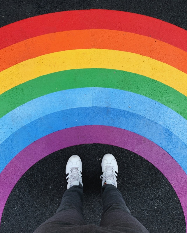 Person standing on a rainbow crosswalk, representing LGBTQ+ pride and visibility.